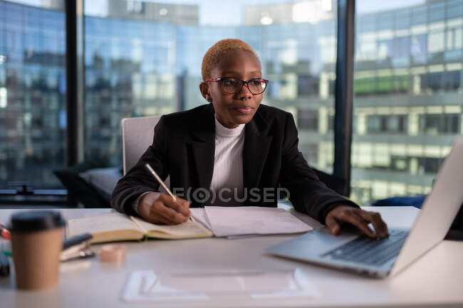 Mulher negra usando laptop e escrevendo em notebook durante o trabalho no escritório moderno — Fotografia de Stock