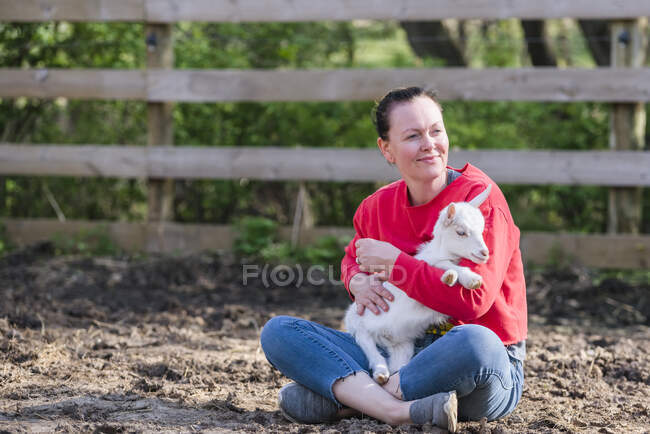 Mujer con sudadera roja sosteniendo una cabra bebé blanca en su regazo. - foto de stock