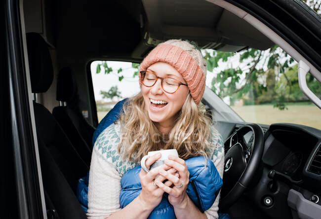 Woman enjoying her cup of coffee laughing whilst sat in her camper van — Stock Photo