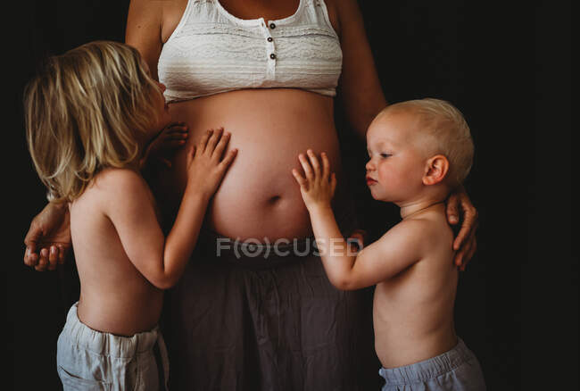 Jóvenes niños felices abrazando tocando el vientre grande embarazada de mamá en casa - foto de stock