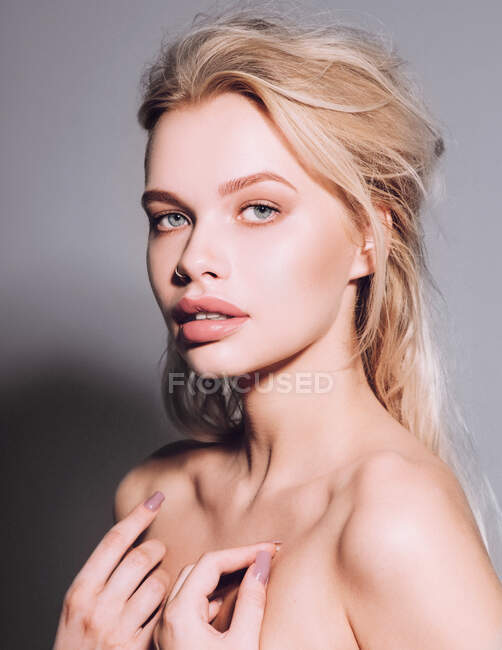 Closeup of pretty young woman face with blue eyes, curly natural blonde hair, has no makeup, touching her soft skin, standing shirtless with bare shoulders, looking at camera. Studio grey background — Stock Photo