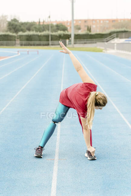 Paralympic professional female runner with leg prosthesis standing at stadium and warming up before training while stretching — Stock Photo