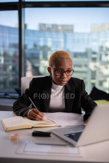 Чорна жінка використовує ноутбук і робить нотатки під час роботи над проектом в офісі — стокове фото