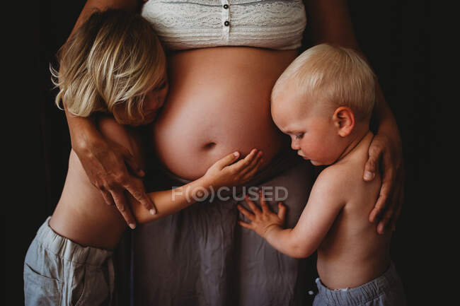 Joven rubia hermanos abrazando mamá embarazada gran vientre en casa - foto de stock