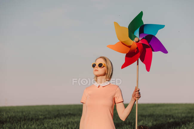 Stile donna in abito rosa con occhiali gialli tenendo girandola su sfondo cielo blu — Foto stock