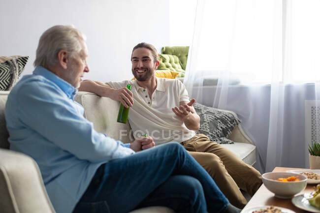 Счастливый молодой человек наслаждается пивом и разговаривает с дедушкой, расслабляясь на диване — стоковое фото