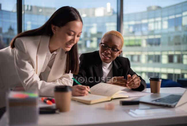 Щасливі багаторасові жіночі колеги посміхаються і пишуть в планувальниці в офісі — стокове фото