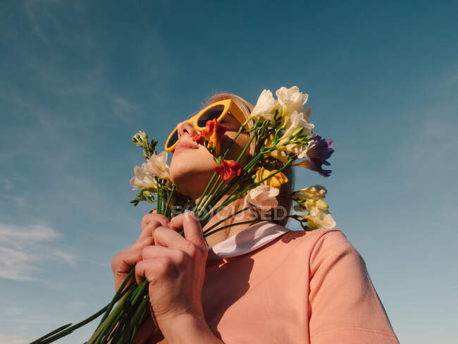 Stile donna in abito rosa con occhiali gialli che tengono fiori su sfondo cielo blu — Foto stock