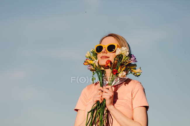 Mujer de estilo en vestido rosa con gafas amarillas sosteniendo flores sobre fondo azul cielo - foto de stock