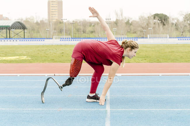 Vue latérale de l'athlète féminine avec une jambe prothétique se penchant vers l'avant et touchant le sol tout en se préparant à courir pendant l'entraînement sur piste et terrain sur le stade — Photo de stock
