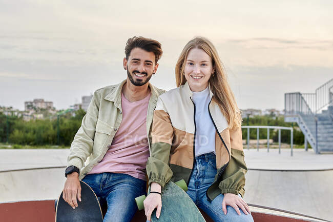 Молодая многонациональная пара смотрит в камеру, улыбаясь в скейт-парке — стоковое фото