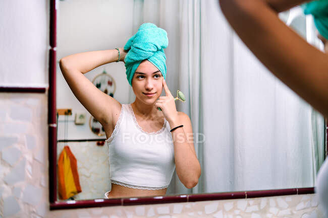 Молода дівчина з рушником на голові робить масаж обличчя у ванній вдома — стокове фото