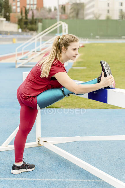 Side view of flexible professional sportswoman with artificial leg stretching legs and leaning on hurdle while preparing for training at stadium — Stock Photo