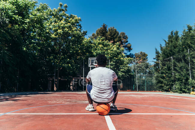 Visão traseira de um menino negro afro sentado em sua bola no campo de basquete. — Fotografia de Stock