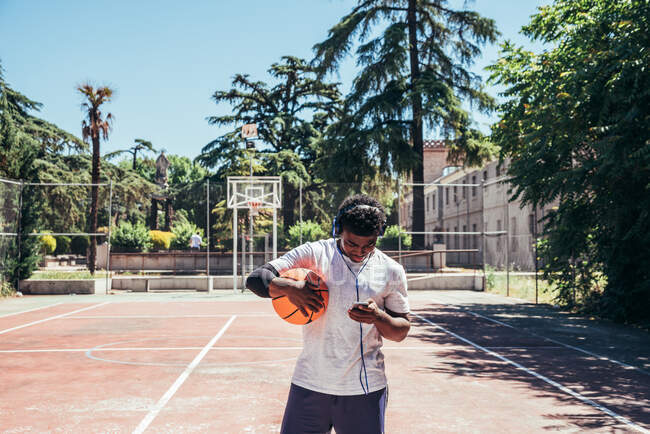 Schwarzer afroamerikanischer Junge hört Musik mit Kopfhörern und seinem Handy und spielt Basketball auf einem städtischen Platz. — Stockfoto
