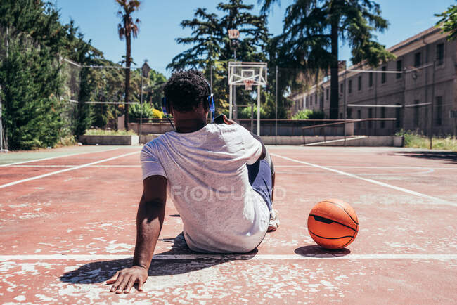 Rückansicht eines schwarzen Afro-Jungen, der sein Handy benutzt und auf dem Basketballplatz mit Kopfhörern Musik hört. Konzepttechnik und Sport. — Stockfoto