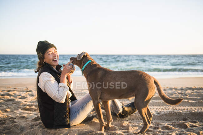 Junge Frau mit ihrem Hund trinkt Kaffee am Strand — Stockfoto