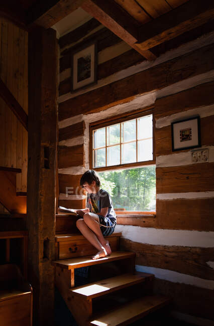 Menino lendo um livro dentro de casa no ensolarado degrau de uma cabana de madeira para casa. — Fotografia de Stock