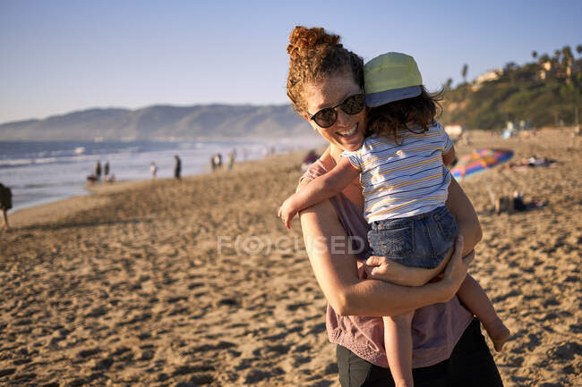 Mère souriante portant sa fille à la plage pendant le week-end — Photo de stock