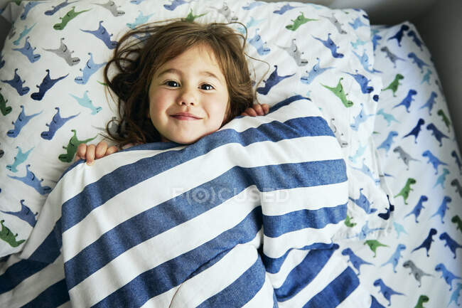 Portrait of cute girl covered with blanket lying on bed at home — Stock Photo