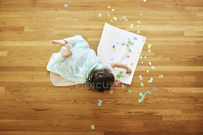 Дівчина робить ремесло з різнокольоровими паперами, лежачи на дерев'яній підлозі вдома — стокове фото