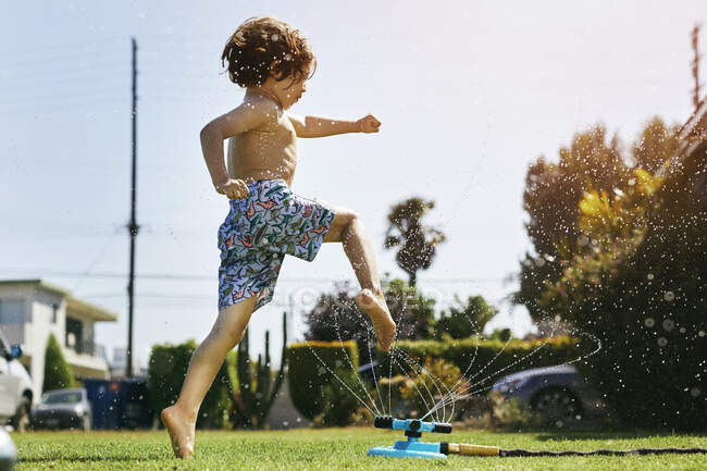 Мальчик без рубашки прыгает через разбрызгиватель на заднем дворе против неба — стоковое фото