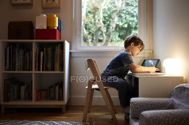 Side view of boy drawing while sitting on chair by window at home — Stock Photo