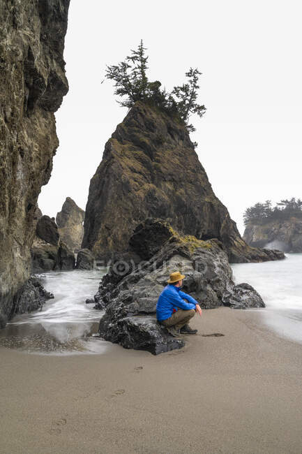 Hombre sentado en la playa con pilas de mar con un abrigo hinchado y sombrero - foto de stock