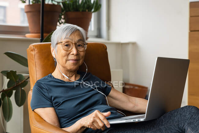 Portrait de femme âgée portant des écouteurs tout en utilisant un ordinateur portable à la maison — Photo de stock