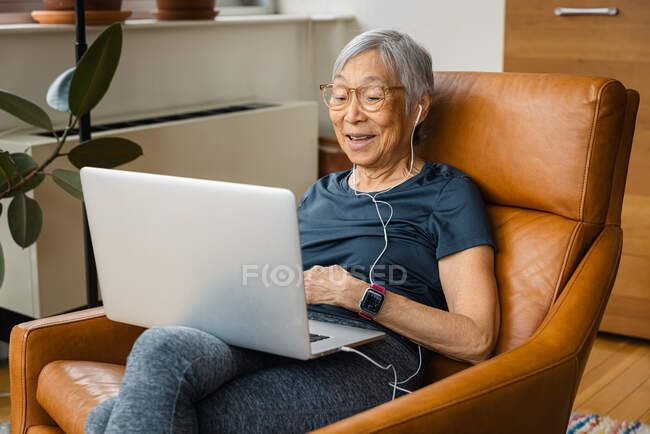 Porträt einer Seniorin, die Kopfhörer trägt, während sie zu Hause ihren Laptop benutzt — Stockfoto