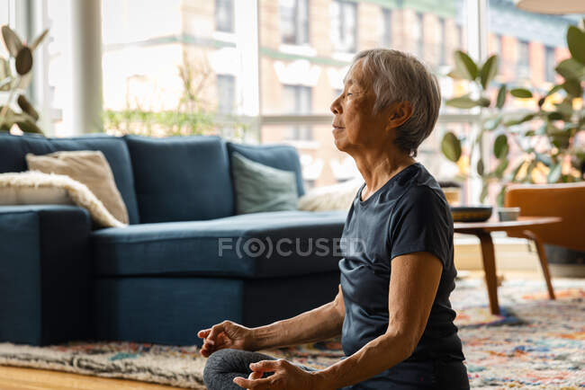 Asiática senior mujer meditando y relajándose en casa en sala de estar - foto de stock