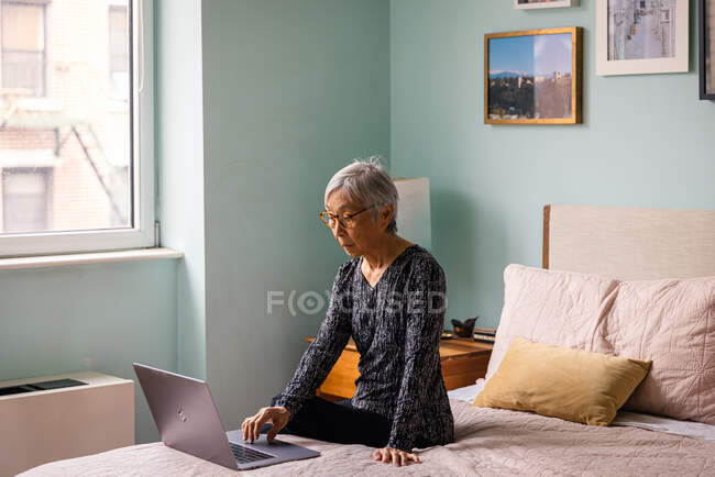 Femme âgée utilisant un ordinateur portable tout en étant assis dans la chambre à coucher à la maison — Photo de stock