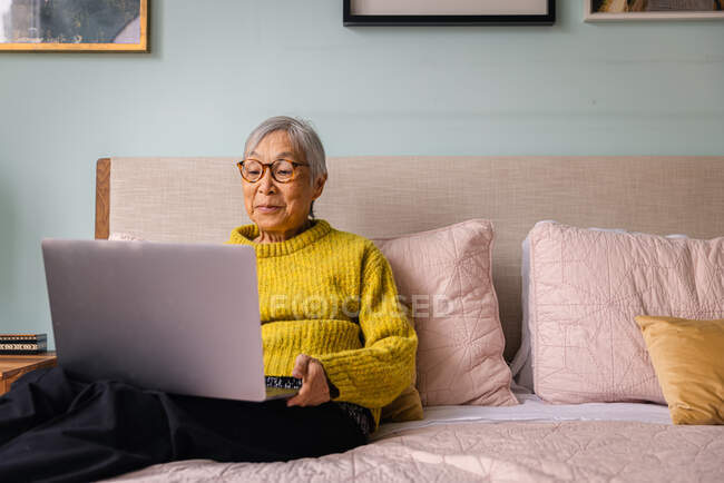 Пожилая женщина использует ноутбук, сидя дома в спальне — стоковое фото