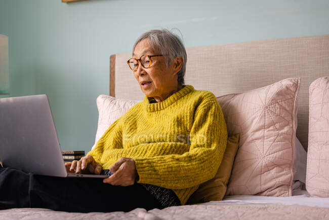 Seniorin benutzt Laptop, während sie zu Hause im Schlafzimmer sitzt — Stockfoto