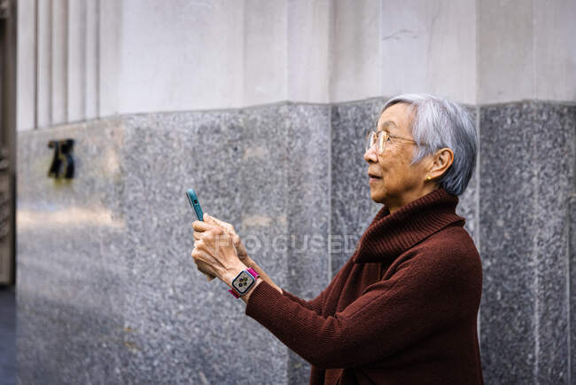 Side view of senior woman using smart phone while standing by wall — Stock Photo