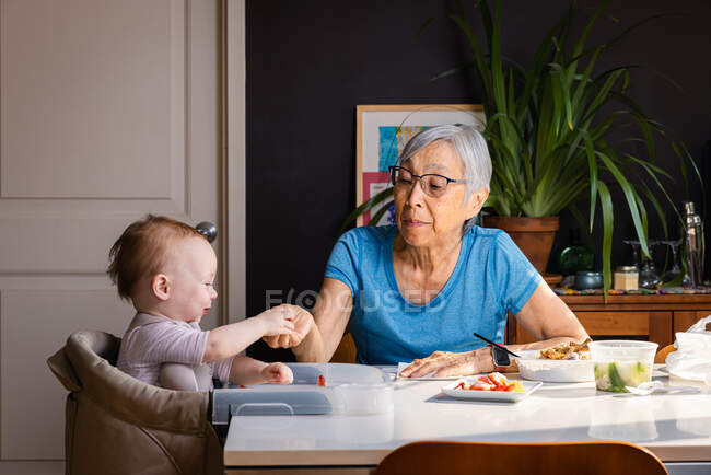 Senior asiatisch frau feeding kleinkind grandaughter at table at home — Stockfoto
