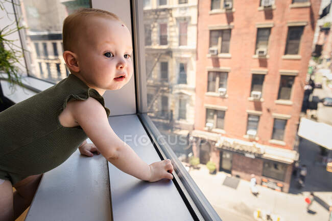 Menina bebê feliz curioso olhando para fora da janela na cidade — Fotografia de Stock