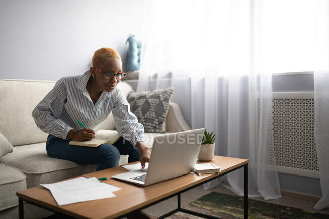 Сучасний молода чорношкіра жінка віддалений працівник в повсякденному одязі використовує ноутбук і робить нотатки в блокноті під час роботи у вітальні вдома — стокове фото