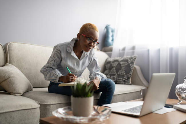 Giovane imprenditrice afroamericana concentrata seduta sul divano e che prende appunti in agenda mentre si incontra online via laptop durante il lavoro remoto da casa — Foto stock