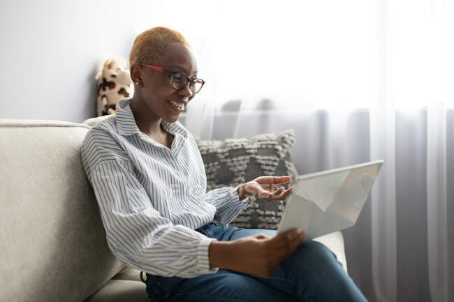 Joven mujer afroamericana positiva en traje casual y gafas sentadas en el sofá y comunicándose con su socio de negocios a través de videollamada en la tableta mientras trabaja de forma remota desde casa - foto de stock