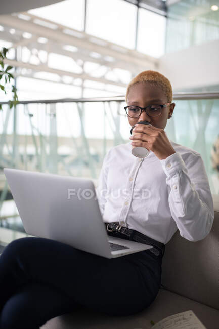 Black female employee sipping coffee to go and reading data on netbook in office — Stock Photo