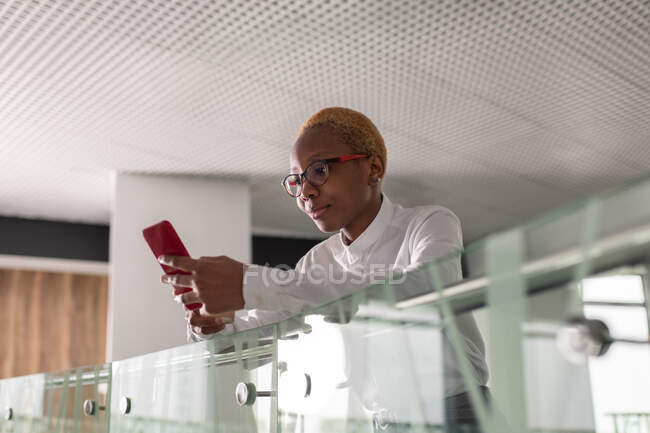Schwarze Verkäuferin lehnt am Geländer und benutzt Handy im Büroflur — Stockfoto