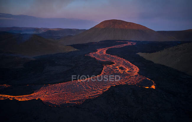 Удивительный горный пейзаж с огненной горячей лавой, протекающей через долину во время извержения вулкана Фаградальсфьолл в Исландии в сумерках — стоковое фото