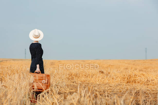 Mujer con sombrero y vestido negro con maleta en el campo de trigo - foto de stock