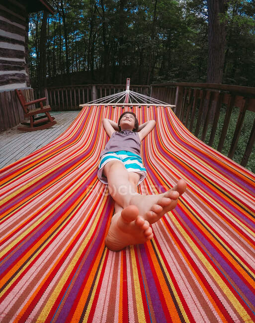 Wide angle of young boy relaxing in a striped hammock on cottage deck. — Stock Photo