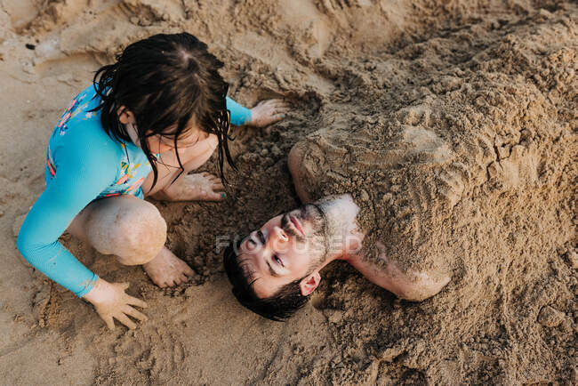 Giovane ragazza seppellisce papà nella sabbia sulla spiaggia di Waikiki durante il tramonto — Foto stock