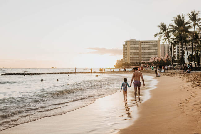 Padre e hija tomados de la mano mientras caminan por la playa al atardecer - foto de stock