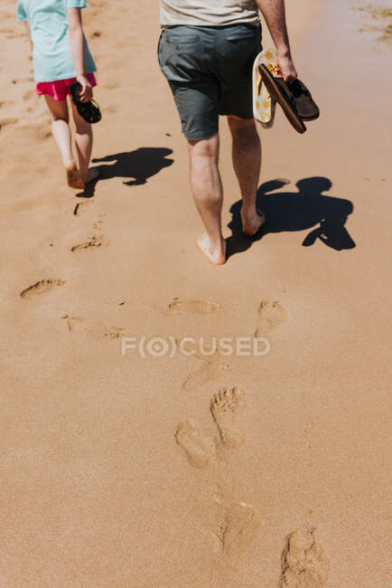 Father and daughter walk across sandy beach while holding flip flops — Stock Photo