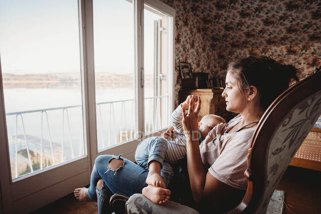 Mother breastfeeding baby on chair in vintage room looking out on lake — Stock Photo