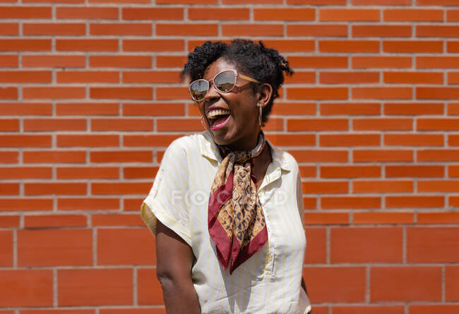Delighted black woman in stylish clothes and sunglasses smiling and looking away while standing against brick wall on street — Stock Photo